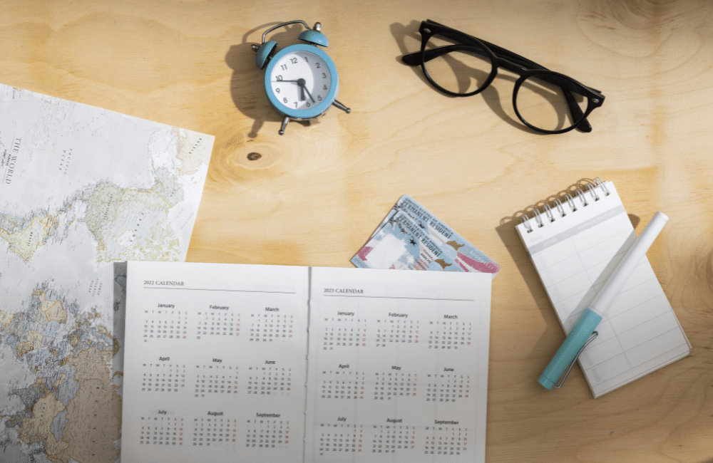 Imagem por freepik | um calendário aberto em cima de uma mesa que está decorada com um óculos de grau, um caderno de anotações e um relógio analógico antigo.